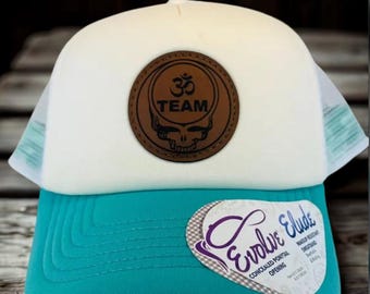 May include: A white and teal trucker hat with a brown leather patch featuring a skull and the text "TEAM" and an Om symbol. The hat has a curved bill and a mesh back. The hat also has a label that says "Evolve Elude".