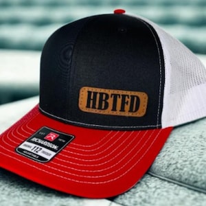May include: A black, red, and white trucker hat with the text "HBTFD" on a brown leather patch. The hat has a red peak, a black front panel, and a white mesh back. The hat is adjustable.