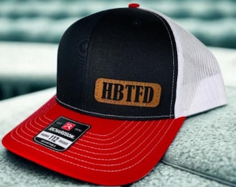 May include: A black, red, and white trucker hat with the text "HBTFD" on a brown leather patch. The hat has a red brim, a black front panel, and a white mesh back. The hat is adjustable.