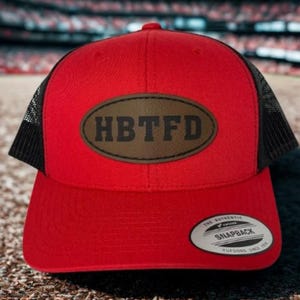 May include: A red trucker hat with a black mesh back and a curved brim. The hat features a brown oval patch with the letters "HBTFD" in black. A white and black Snapback tag is on the front.