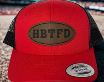 May include: A red trucker hat with a black mesh back and a curved brim. The hat features a brown oval patch with the letters "HBTFD" in black. A white and black Snapback tag is on the front.
