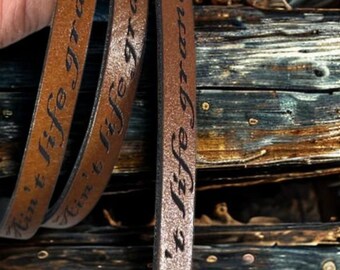 May include: Three brown leather bracelets with the phrase "Ain't life grand" engraved in black. The bracelets are held against a rustic wooden background, showcasing the texture and color of the leather.