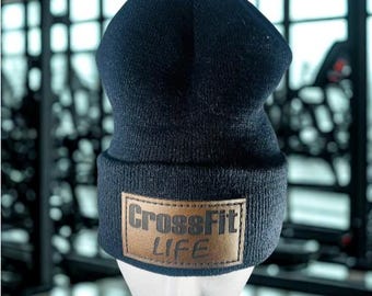 May include: A dark navy blue knit beanie with a brown leather patch. The patch features the words "CrossFit LIFE" in a bold, textured font. The beanie is displayed on a white mannequin head, set against a blurred gym background.