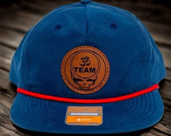 May include: A blue baseball cap with a red rope detail and a brown leather patch. The patch features a skull design with the word "TEAM" and an Om symbol. The cap has a flat brim and a Richardson tag.