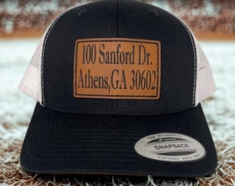 May include: Black and white trucker hat with a brown leather patch. The patch is embossed with the address "100 Sanford Dr. Athens, GA 30602". The hat has a snapback closure and a Yupoong label.