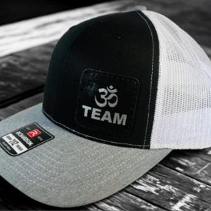 Puede incluir: Gorra de camionero negra y blanca con visera gris. La gorra presenta un parche con el símbolo Om y la palabra "TEAM" en plata. La etiqueta Richardson 112 Trucker es visible en la visera.