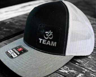 May include: A black and white trucker hat with a gray brim. The hat features a patch with the Om symbol and the word "TEAM" in silver. The Richardson 112 Trucker tag is visible on the brim.