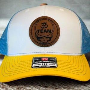 May include: A white and blue trucker hat with a yellow peak. The hat features a brown leather patch with the text "TEAM" and a skull design. The hat is adjustable and has a Richardson 112 tag.