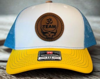 May include: A white and blue trucker hat with a yellow brim. The hat features a brown leather patch with the text "TEAM" and a skull design. The hat is adjustable and has a Richardson 112 tag.