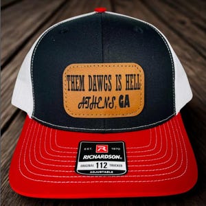 May include: A black, white, and red trucker hat with the text "THEM DAWGS IS HELL ATHENS, GA" on a leather patch. The hat has a red brim, a black crown, and a white mesh back. The Richardson brand label is visible.