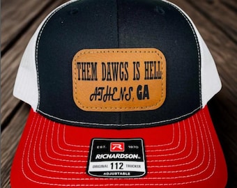 May include: A black, white, and red trucker hat with the text "THEM DAWGS IS HELL ATHENS, GA" on a leather patch. The hat has a red brim, a black crown, and a white mesh back. The Richardson brand label is visible.