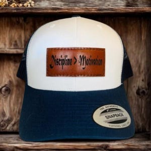 May include: A white and navy blue snapback hat with a brown leather patch that reads "Discipline > Motivation". The hat has a mesh back and a curved brim. The hat has a snap closure.