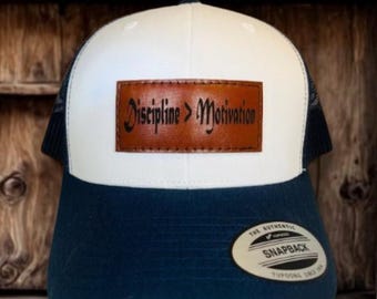 May include: A white and navy blue snapback hat with a brown leather patch that reads "Discipline > Motivation". The hat has a mesh back and a curved brim. The hat has a snap closure.