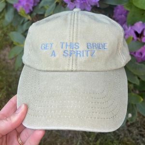 May include: A light green baseball cap with the text "GET THIS BRIDE A SPRITZ" embroidered in light blue. The cap is made of a soft fabric and has a curved brim. The background features purple flowers and green foliage.