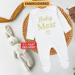 May include: White baby onesie with long sleeves and gold embroidery that reads "Baby Mear" with a heart.