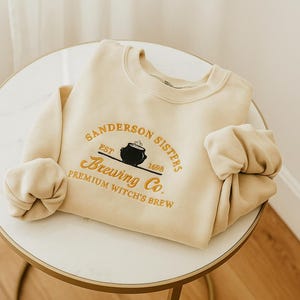May include: Cream-colored sweatshirt with the text "Sanderson Sisters Brewing Co." in gold lettering. The design includes a black cauldron and the text "Premium Witch's Brew". The sweatshirt is folded on a white table.