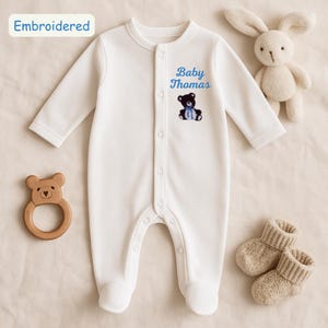 Peut inclure: Combinaison blanche pour bébé à manches longues et boutons pression. La combinaison a le texte "Baby Thomas" brodé sur le devant, ainsi qu'un petit ours en peluche noir brodé portant un nœud papillon bleu.