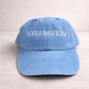Może przedstawiać: Jasnoniebieska czapka bejsbolówka z haftowaną na przodzie białą datą zapisaną rzymskimi cyframi "XXII.II.MMXXV". Czapka ma zakrzywiony daszek i regulowany pasek. Wykonana z miękkiego, pranego materiału bawełnianego.