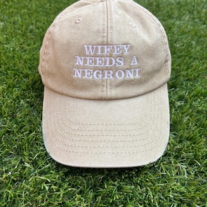 May include: A tan baseball cap with the embroidered text "Wifey Needs A Negroni" in white. The cap has a curved brim and a classic six-panel design. The hat is made of a durable fabric.