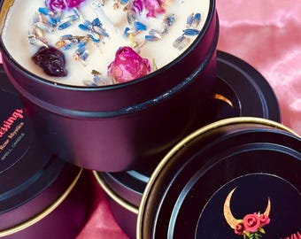 May include: Black candle tins with gold rims, filled with cream-colored wax, dried lavender, and rose buds. The tins are labeled "Sacred Blessings" with a crescent moon and floral design. The candles are on a pink surface.