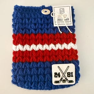 May include: Handmade blue, red, and white knitted Kindle cover. The cover features a button closure and a patch with crossed hockey sticks and the numbers 24 and 81. A tag reads "Kindle Cover".