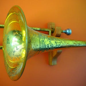 May include: A vintage brass trombone with a tarnished finish, mounted on a wooden holder against an orange background. The instrument features a bell and slide, with visible text on the bell. A classic musical instrument.