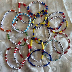 May include: A collection of beaded bracelets in various colors, spelling out country names like Argentina, Austria, and Ecuador. The bracelets feature white letter beads and colorful accents, including red, blue, yellow, and green beads. The bracelets are displayed on a white, textured surface.