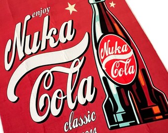 May include: A rectangular red towel featuring a graphic of a Nuka-Cola bottle with the text "enjoy Nuka Cola classic original 2044" and "ICE COLD" printed in white and black lettering, accented by white stars on a solid red background.