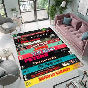 Puede incluir: Una alfombra colorida con una pila de cintas VHS con títulos de películas de terror, incluyendo "La Maldición de Amityville", "Chucky", "Frankenstein", "Candyman", "Tiburón", "Los Pájaros", "Psicosis", "Halloween", "Re-Animator", "El silencio de los corderos" y "El día de los muertos".