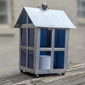 May include: A decorative metal lantern shaped like a small house, with a silver roof and frame. The windows are filled with blue glass. A small bird ornament sits on top. A white candle is inside.