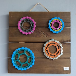 May include: A wooden wall hanging featuring four colorful bicycle chainrings. The chainrings are painted in shades of pink, blue, orange, and yellow, and are mounted on a dark brown wooden plank. The piece is suspended by a natural rope.