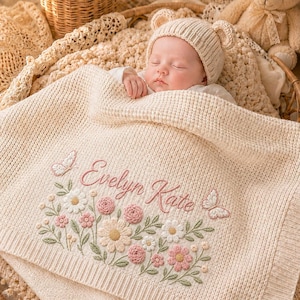 May include: A cream-colored knit baby blanket featuring embroidered pink flowers, green leaves, and two butterflies, with the name Evelyn Kate stitched in pink script. The blanket is draped over a baby wearing a matching cream-colored bear-ear hat.
