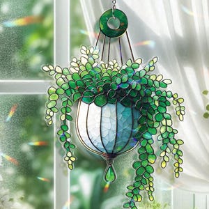 May include: A stained glass hanging planter with a white and blue pot, filled with green leafy plants. The planter is suspended by a chain and ring, with a green teardrop-shaped charm at the bottom. The background includes a window and curtains.
