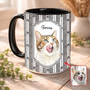 May include: A ceramic mug with a black handle and interior. The mug features a portrait of a cat with its tongue out, labeled "Bonnie" within a decorative frame. The background has gray and white stripes with bow accents.