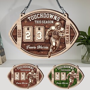 Könnte beinhalten: Holzschild in Footballform mit dem Text "TOUCHDOWNS THIS SEASON" und einer Footballspieler-Grafik. Das Schild zeigt die Zahlen "23" und den Namen "Travis Morris". Inklusive Kette zum Aufhängen.
