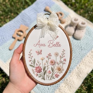 May include: An oval wooden embroidery hoop containing white fabric with the embroidered name Ava Belle in pink script. The design features pink and white wildflowers, green stems, and a small butterfly. A white lace bow is attached to the top.
