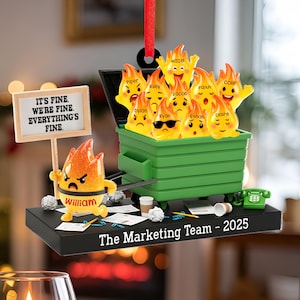 May include: A humorous ornament featuring a green dumpster overflowing with cartoon flames, each with a name. A smaller flame figure holds a sign that reads "IT'S FINE. WE'RE FINE. EVERYTHING'S FINE." The base reads "The Marketing Team - 2025."