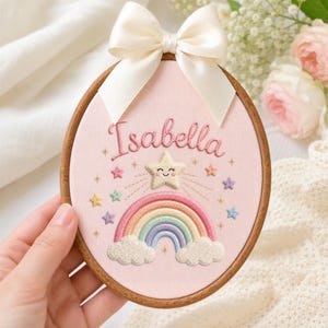 May include: An oval wooden embroidery hoop containing pink fabric with the embroidered name Isabella in cursive script. Below the name is a smiling star, a multi-colored rainbow, and two clouds, surrounded by small embroidered stars and a cream bow.
