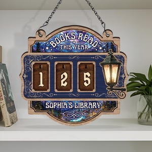 May include: Wooden sign with the text "BOOKS READ THIS YEAR" above a counter displaying "125". The sign has a blue background with a decorative border and a lantern on the right side. The text "SOPHIA'S LIBRARY" is at the bottom.