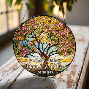 May include: A round stained glass piece featuring a tree with pink and yellow leaves, blue sky, and a brown trunk. The text reads "50th Anniversary David & Bella 07.28.1975". The piece is hanging from a chain.