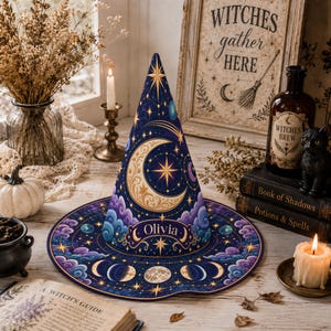 May include: A conical witch hat featuring a deep blue background with gold celestial motifs, including a large crescent moon, stars, and a moon phase cycle. The hat displays the name Olivia in white text and is decorated with purple clouds.