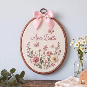 May include: An oval wooden embroidery hoop containing cream-colored fabric with hand-stitched floral designs in pink, white, and green. The name Ava Belle is embroidered in pink script, accompanied by two small butterflies and a pink satin bow.