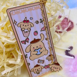 Puede incluir: Un brazalete de charms de oro con un charm de esmalte rosa y blanco con un perro de dibujos animados sosteniendo una galleta. El charm est&aacute; unido a una cadena de oro con tres charms m&aacute;s peque&ntilde;os del mismo perro en diferentes poses.