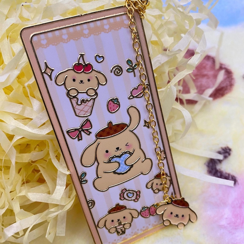 Puede incluir: Un brazalete de charms de oro con un charm de esmalte rosa y blanco con un perro de dibujos animados sosteniendo una galleta. El charm est&aacute; unido a una cadena de oro con tres charms m&aacute;s peque&ntilde;os del mismo perro en diferentes poses.