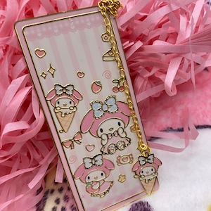 Puede incluir: Un marcap&aacute;ginas rosa y dorado con una cadena y un charm con My Melody, un personaje japon&eacute;s popular. El marcap&aacute;ginas tiene un fondo a rayas rosa y blanco con un coraz&oacute;n rosa y una estrella dorada. El charm es un peque&ntilde;o cono de helado rosa My Melody.