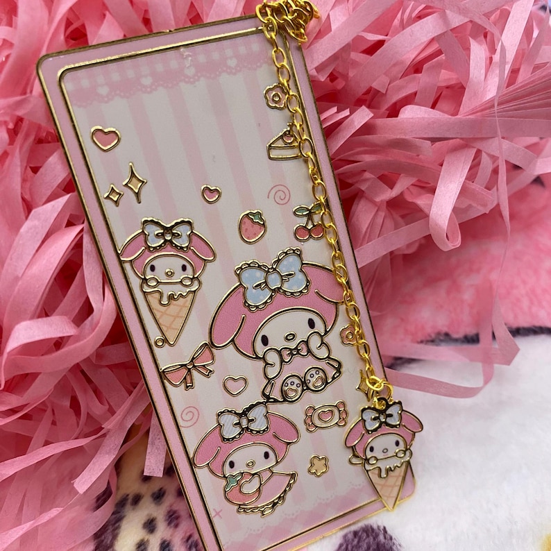 Puede incluir: Un marcap&aacute;ginas rosa y dorado con una cadena y un charm con My Melody, un personaje japon&eacute;s popular. El marcap&aacute;ginas tiene un fondo a rayas rosa y blanco con un coraz&oacute;n rosa y una estrella dorada. El charm es un peque&ntilde;o cono de helado rosa My Melody.