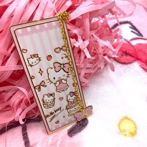 Puede incluir: Un marcap&aacute;ginas de metal dorado con un fondo a rayas rosa y blanco. El marcap&aacute;ginas presenta a Hello Kitty en varias poses y el texto "Hello Kitty". Una cadena dorada conecta el marcap&aacute;ginas a un amuleto de mariposa rosa.