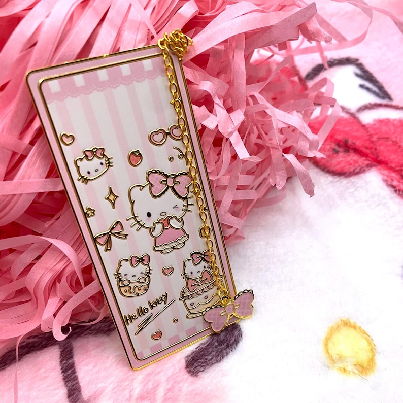 Puede incluir: Un marcap&aacute;ginas de metal dorado con un fondo a rayas rosa y blanco. El marcap&aacute;ginas presenta a Hello Kitty en varias poses y el texto "Hello Kitty". Una cadena dorada conecta el marcap&aacute;ginas a un amuleto de mariposa rosa.