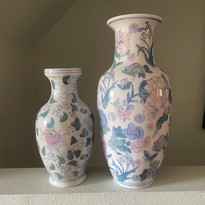 Może przedstawiać: Dwa białe ceramiczne wazony z kwiatowymi wzorami. Większy wazon ma różowe, niebieskie i zielone kwiaty. Mniejszy wazon ma różowe, zielone i niebieskie kwiaty.