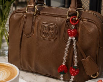 May include: A brown leather handbag with a decorative strawberry charm. The bag has a top handle and a logo embossed on the front. A cup of coffee and a plate of strawberries are in the foreground.